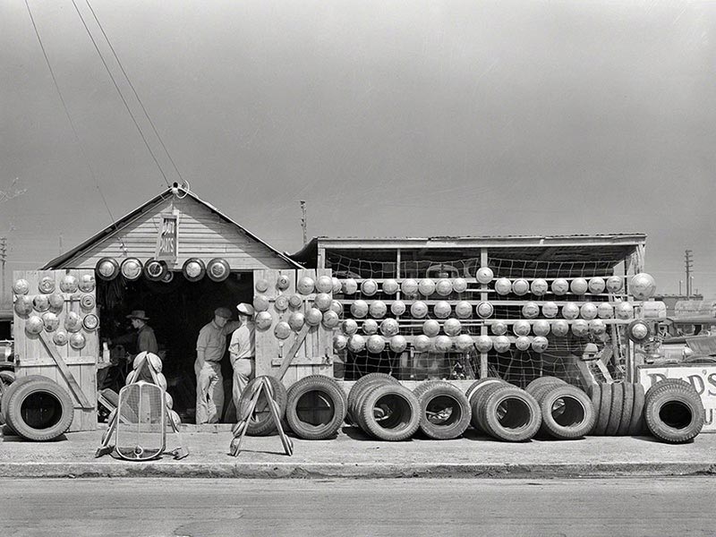 A small roadside shop with tires lined up front and rows of hubcaps hanging on the wall, with a few people standing inside.